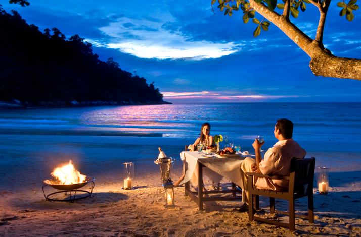 Romantic couple having beachside dinner at sunset, with a fire pit and ocean view at Pangkor Laut Resort.
