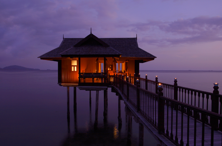 Overwater villa lit at twilight, with a wooden walkway extending over calm sea at Pangkor Laut Resort.