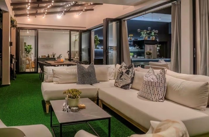 Modern penthouse lounge with cream sofas, decorative pillows, and a green artificial grass floor, opening onto a dining area and kitchen.