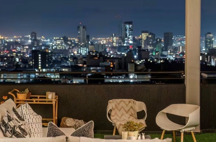 Outdoor seating area on a penthouse balcony at night, with city lights and skyscrapers visible in the distance.
