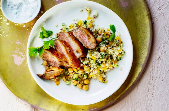 Plate of sliced grilled meat served with chickpeas and a colorful grain salad, garnished with fresh herbs.