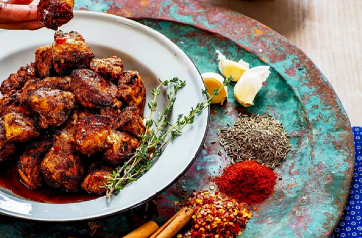 Bowl of spiced grilled meat pieces, surrounded by fresh herbs, garlic, and assorted spices on a rustic tray.