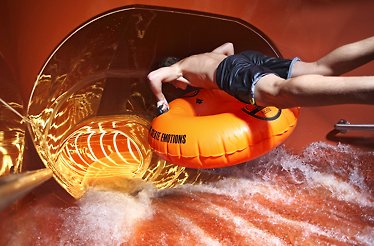 A young man on an inflatable ring speeds through a glowing water slide, warm light reflecting off swirling water as he leans forward in a moment of pure adrenaline.