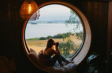 A peaceful retreat framed by a large circular window overlooking calm water and open fields, where soft evening light, warm wooden interiors, and a quiet cup in hand create a feeling of stillness, comfort, and escape from the outside world.