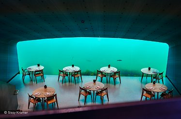 A serene underwater dining room with soft blue light, where guests dine beside a vast ocean view and drifting fish. Photo: Sissy Kramer