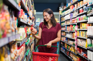 Persona en un pasillo de supermercado sosteniendo un producto para revisar su etiqueta mientras empuja una canasta de compras. A ambos lados hay estanterías llenas de alimentos y productos de consumo.