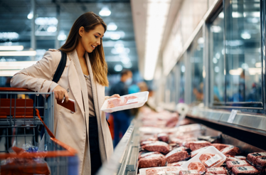 Persona en la sección de carnes de un supermercado revisando la etiqueta de una bandeja de producto refrigerado. A su lado hay un carro de compras y vitrinas con distintos cortes de carne en exhibición.