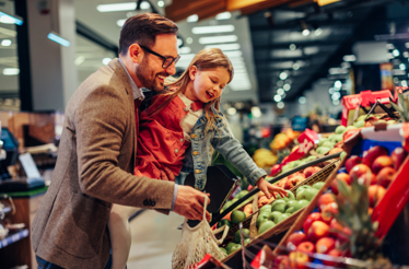 Persona sosteniendo una bolsa reutilizable mientras acompaña a otra persona a elegir frutas y verduras en la sección de productos frescos de un supermercado. Los exhibidores contienen variedad de frutas organizadas en compartimentos.