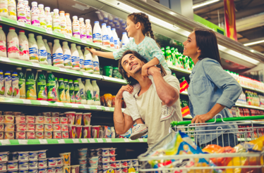 Personas comprando en el pasillo de productos lácteos de un supermercado. Una persona alcanza artículos en un estante mientras otra empuja un carro de compras lleno de productos frente a góndolas refrigeradas.