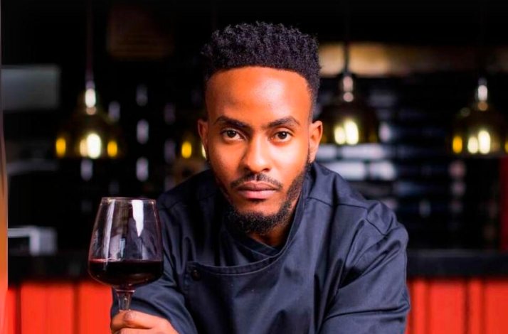 Chef in a dark uniform holding a glass of red wine at a warmly lit restaurant, for Nairobi Supper Club.