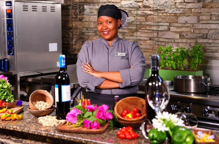  Female chef in grey uniform standing arms crossed in a professional kitchen with wine, herbs, and fresh produce.