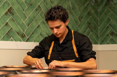 A chef in a black shirt and apron carefully plating dishes at a counter against a green herringbone-tiled wall.