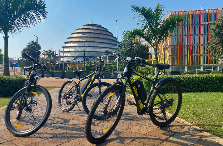 Two 'ebee' e-bikes parked on a sunny Kigali plaza, with the iconic Kigali Convention Centre dome and colorful building visible behind.