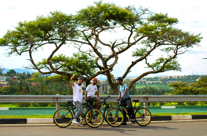 Three cyclists pausing for a selfie with black-and-yellow e-bikes on a Kigali road, beneath a wide acacia tree with hilly cityscape behind.