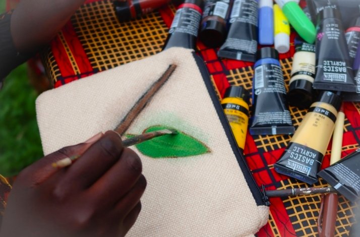 Person painting a green leaf on a fabric pouch, surrounded by art supplies.