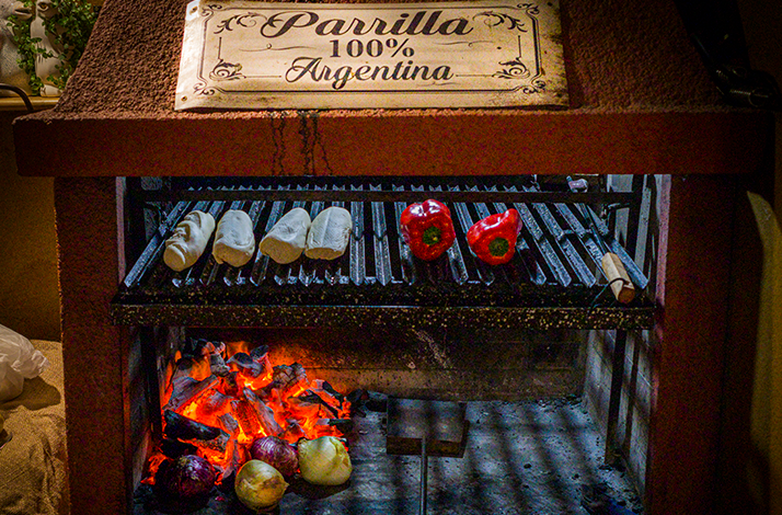 An Argentine grill cooking bread and red bell peppers over hot coals under a Parrilla 100% Argentina sign.