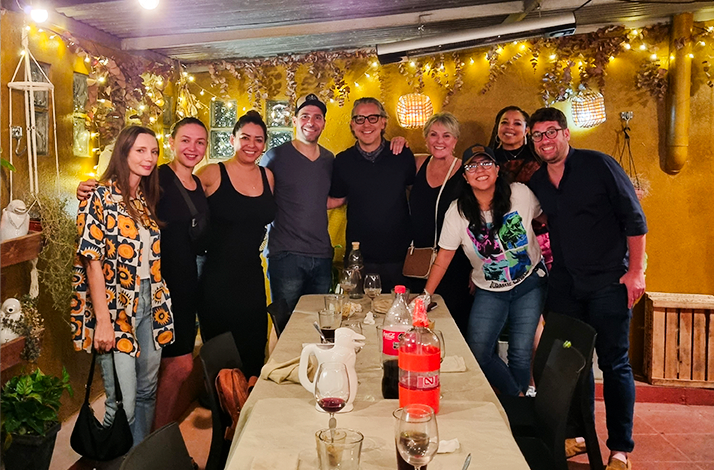 A group of smiling friends poses behind a long dinner table under warm string lights on an outdoor patio.