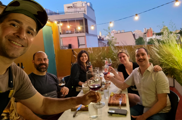 A group of friends smiling and raising wine glasses for a toast on an outdoor rooftop patio at dusk.