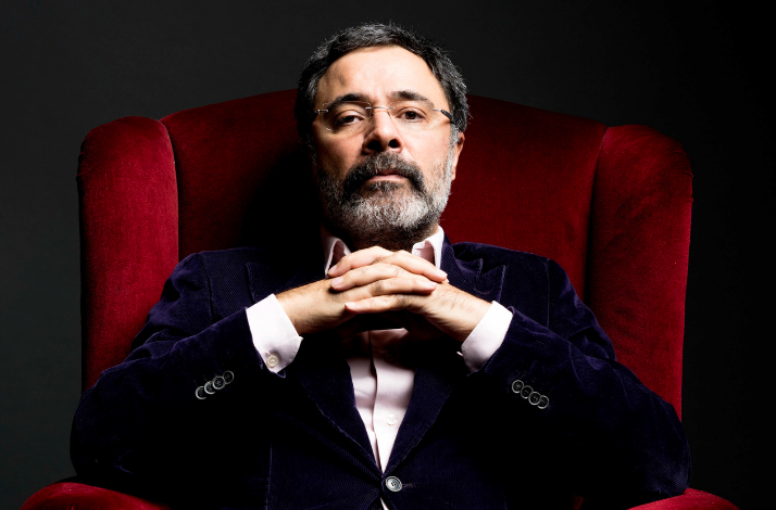 A portrait of author Ahmet Ümit sitting in a red velvet armchair against a dark background. He has grey hair and a beard, wears glasses and a dark blazer, and has his hands clasped under his chin, looking directly at the camera.