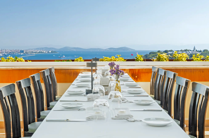 A perspective view of a long dining table set with white linens, plates, glassware, and small purple flowers. In the background, the blue waters of the Bosphorus and the silhouette of Istanbul's Historic Peninsula are visible under bright daylight.