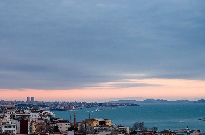 A wide-angle cityscape of the Bosphorus in Istanbul. Under a slightly cloudy sky with a hint of sunset glow, the sea is surrounded by city buildings, mosque minarets, and distant islands. A ferry is seen traveling across the water.