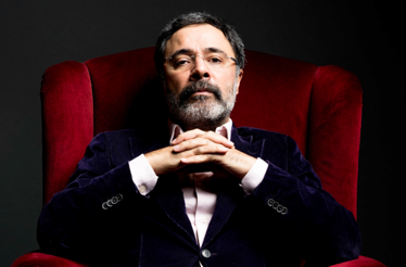 A portrait of author Ahmet Ümit sitting in a red velvet armchair against a dark background. He has grey hair and a beard, wears glasses and a dark blazer, and has his hands clasped under his chin, looking directly at the camera.
