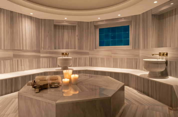 A serene and modern Turkish hammam featuring grey-veined marble walls, an octagonal central massage stone with burning candles, and traditional basins.