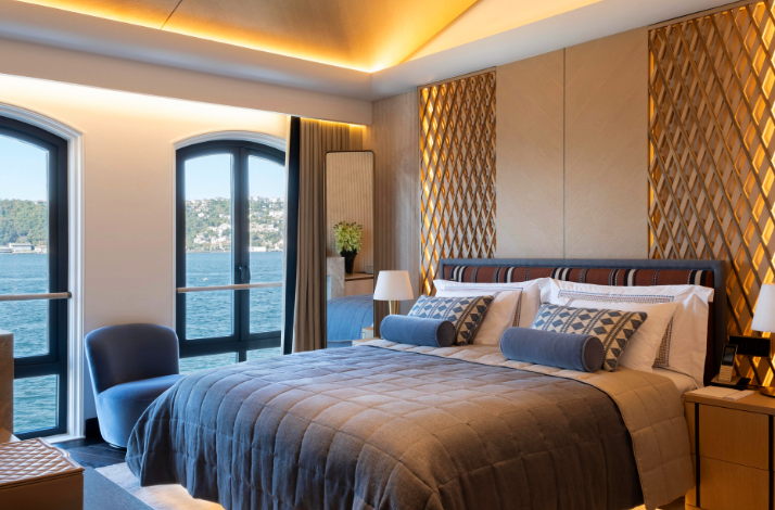 An elegant hotel room featuring a comfortable double bed with blue accents, geometric wooden wall panels, and large arched windows offering a stunning view of the Bosphorus.