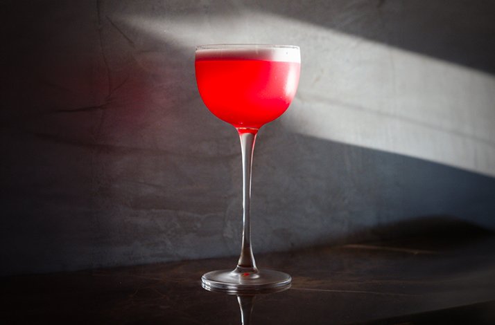 Vibrant crimson cocktail with a frothy white foam top served in a tall coupe glass at Alo Restaurant.