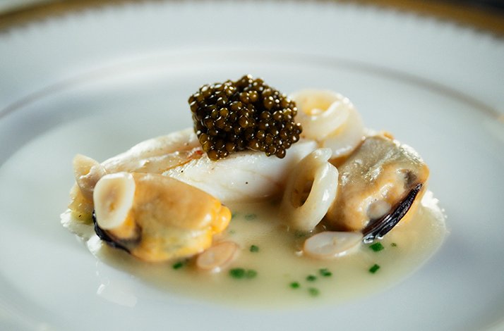 Seared fish fillet topped with caviar, served alongside oysters and clams in a delicate broth at Alo Restaurant.