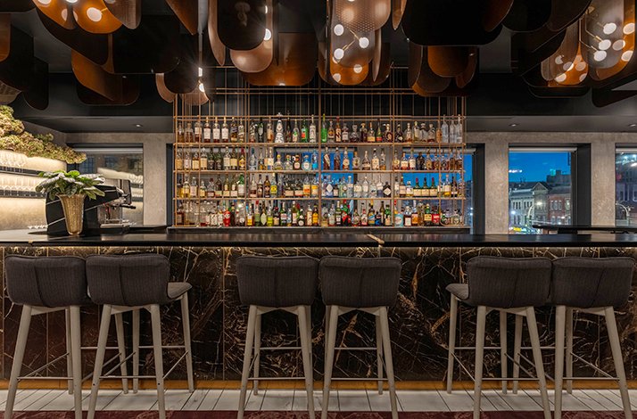Dark marble bar at Alo Restaurant featuring copper ceiling fixtures and a floor-to-ceiling bottle display with city views.