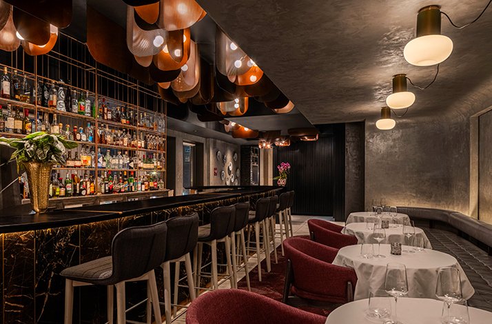 Alo Restaurant's dining room featuring a marble bar, copper ceiling installation, burgundy chairs, and white-clothed tables.