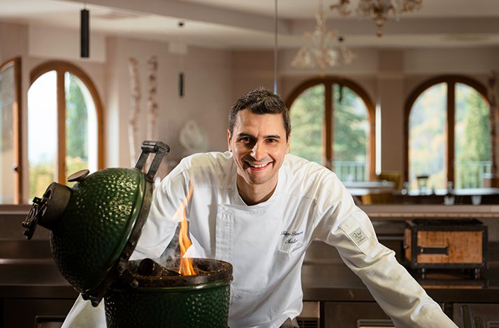 Chef Filippo Baroni in white uniform standing behind a green kamado grill with an open flame.