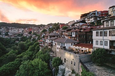 Panoramic sunset view over Veliko Tarnovo's hillside old town, with terracotta rooftops, stone cliffs, lush greenery, and a glowing pink sky.