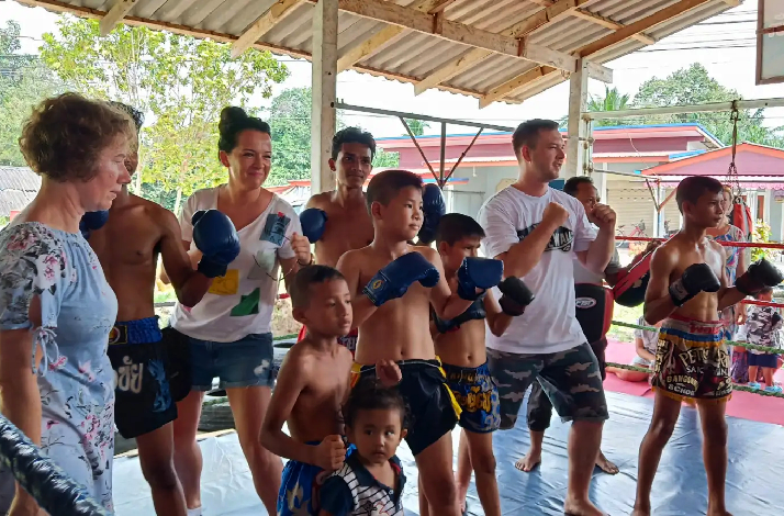 Group of adults and children posing together in a Muay Thai boxing gym, wearing boxing gloves and shorts.