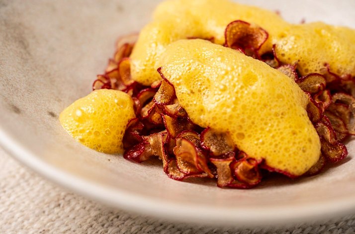 Plated dish at Afluente in Bogota, Colombia, featuring thinly sliced red-rimmed root vegetable crisps topped with an aerated yellow foam, served in a speckled ceramic bowl