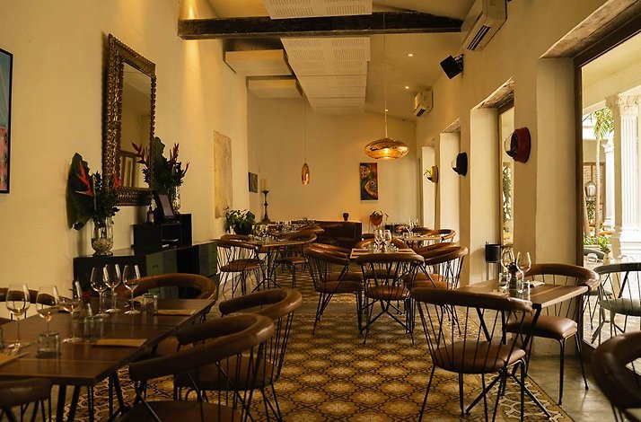 Interior dining room at Carmen, Cartagena, Colombia: leather-backed chairs and set tables with wine glasses on patterned hydraulic-tile floors, a copper pendant lamp and gilt mirror, with an archway opening to a planted courtyard.