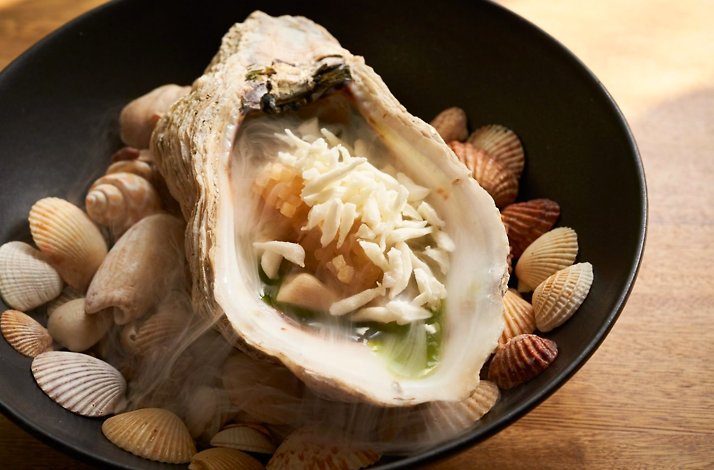 Plated seafood dish at Carmen, Cartagena: an oyster served in its shell over a bed of small shells in a black bowl with a smoky presentation.