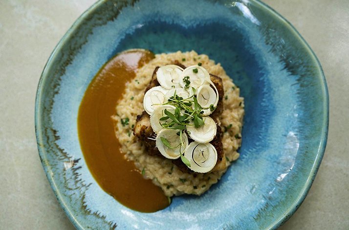 Plated main course at Carmen, Cartagena: a crisp-topped protein over creamy risotto with jus, garnished with thin radish slices and microgreens, in a blue glazed ceramic bowl.