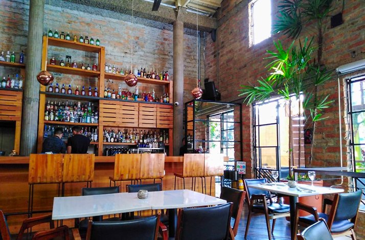 Dining room at Carmen, Medellin: exposed-brick walls, a wood bar lined with bottles under copper pendant lamps, set tables with white tops and dark chairs, and tall green plants by large windows