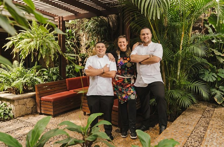 Carmen - Three members of the kitchen team standing arm in arm in a lush tropical garden patio with ferns and palms; two wear white chef jackets and one a colorful floral apron, beside a wooden pergola.