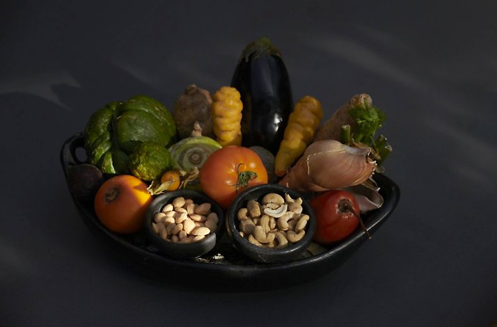 A dark ceramic dish of fresh Colombian ingredients — eggplant, tomatoes, avocados, native tubers, beets and small bowls of peanuts and cashews, dramatically lit against a dark backdrop, at El Chato restaurant in Bogota, Colombia
