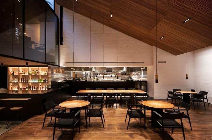 Interior dining room at Restaurante Leo in Bogotá, Colombia, with a vaulted wood-slat ceiling, wood floors, dark chairs around round wood tables, and an illuminated open bar and kitchen pass at the rear.