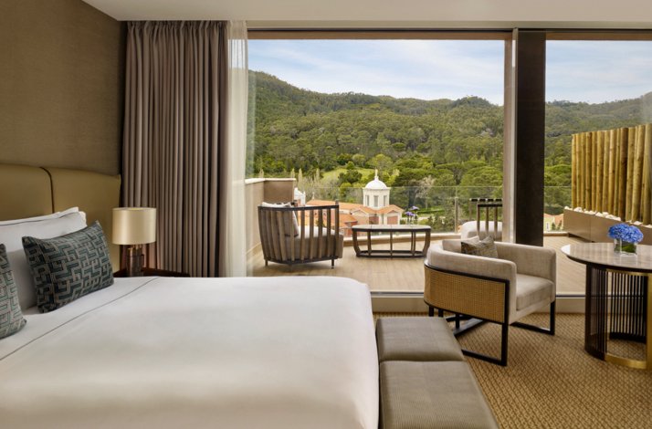 Elegant hotel room at Penha Longa Resort, containing a large bed and seating area, opening onto a balcony with scenic views of forested hills and a historic chapel.