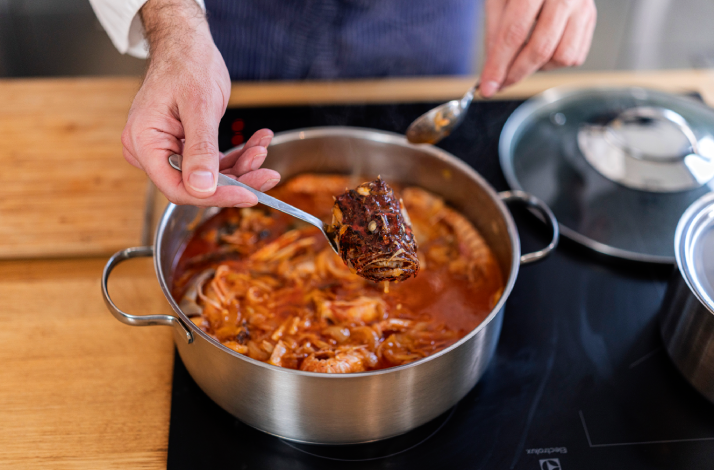 Chef lifting a braised fish piece with tongs from a simmering Dalmatian brudet stew on an induction hob.