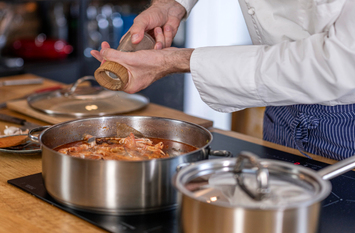 Chef in white coat seasoning Dalmatian brudet with a wooden pepper mill over a steel pan on an induction hob.