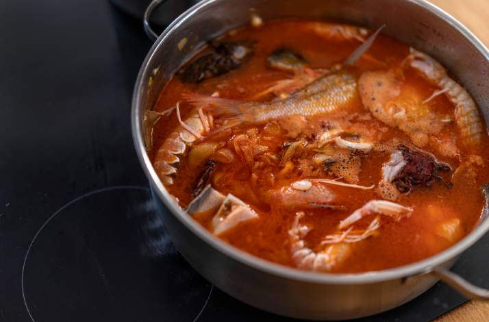 Mixed seafood brudet stew bubbling in a steel pot on an induction hob, featuring prawns, fish, and squid.
