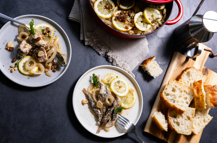 Two plated servings of Dalmatian marinated fish with lemon, onions, and pine nuts, with a red pot and bread board alongside.