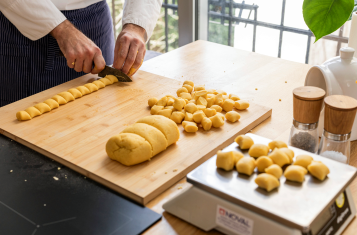 Chef slicing gnocchi dough rope into portions on a wooden board, with shaped pieces and a kitchen scale alongside.
