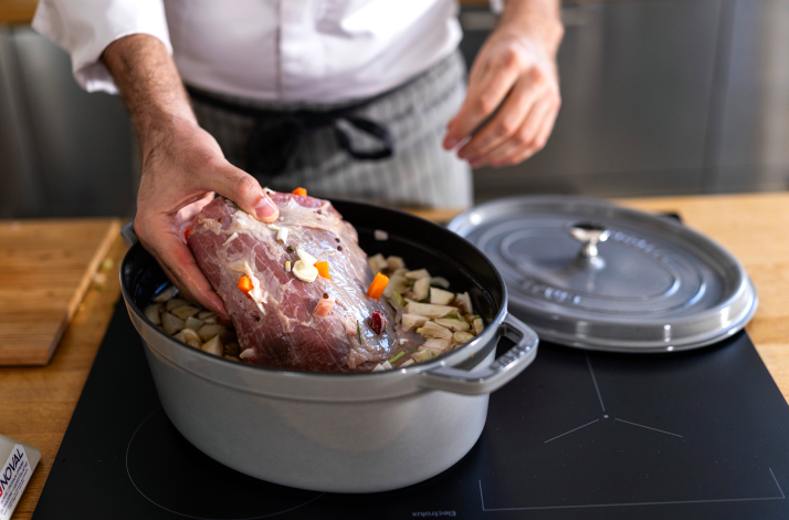 Chef lowering a larded raw beef joint into a cast-iron pot over chopped vegetables, ready for braising.